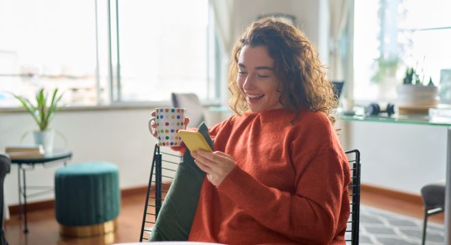 Junge lächelnde Frau hält Smartphone und trinkt zu Hause entspannt Kaffee.