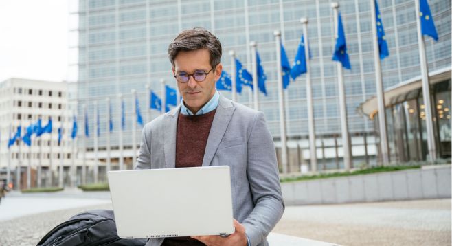 Geschäftsmann arbeitet an einem Laptop vor dem Gebäude der Europäischen Kommission mit mehreren EU-Flaggen im Hintergrund