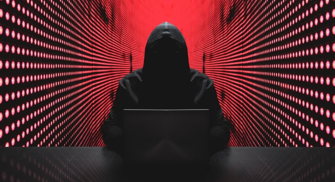 In Dunkelheit gehüllter Hacker sitzt vor einem Laptop, der Hintergrund bedrohlich rot