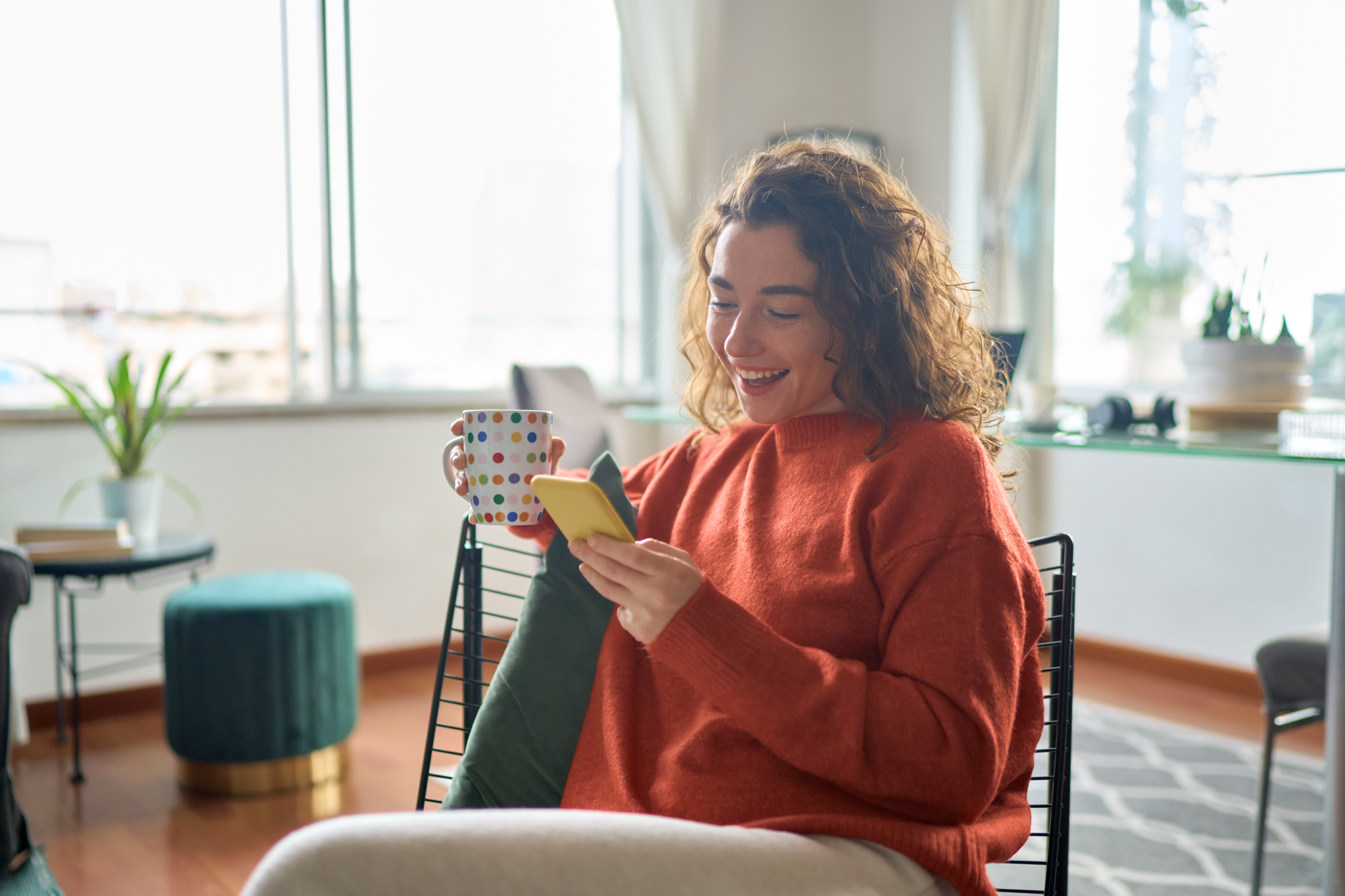 Young woman sits with coffee in the living room and looks at her mobile phone with a smile