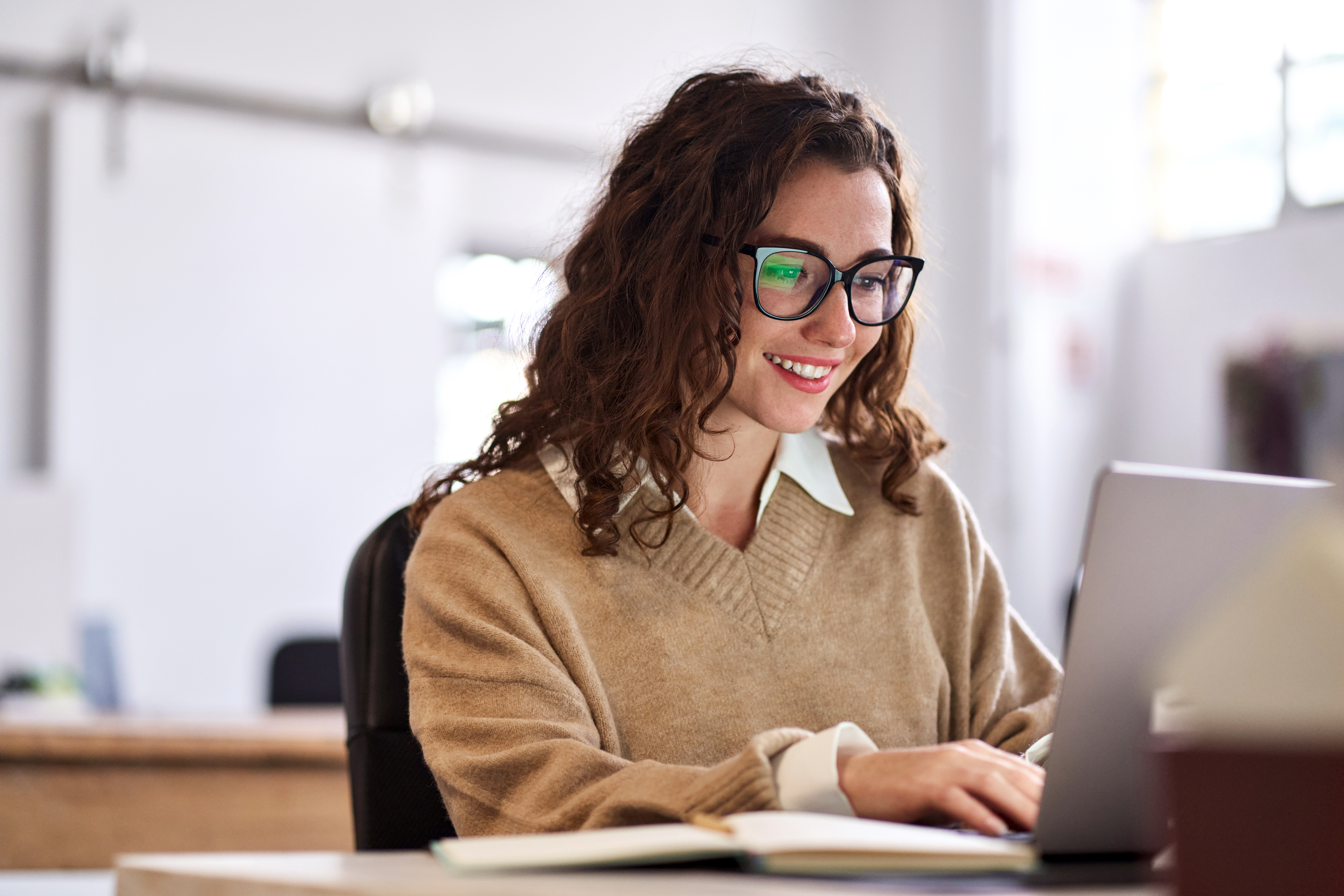 A person with curly hair sits at a desk, focused on a laptop, wearing a cozy beige sweater in a bright, modern workspace.