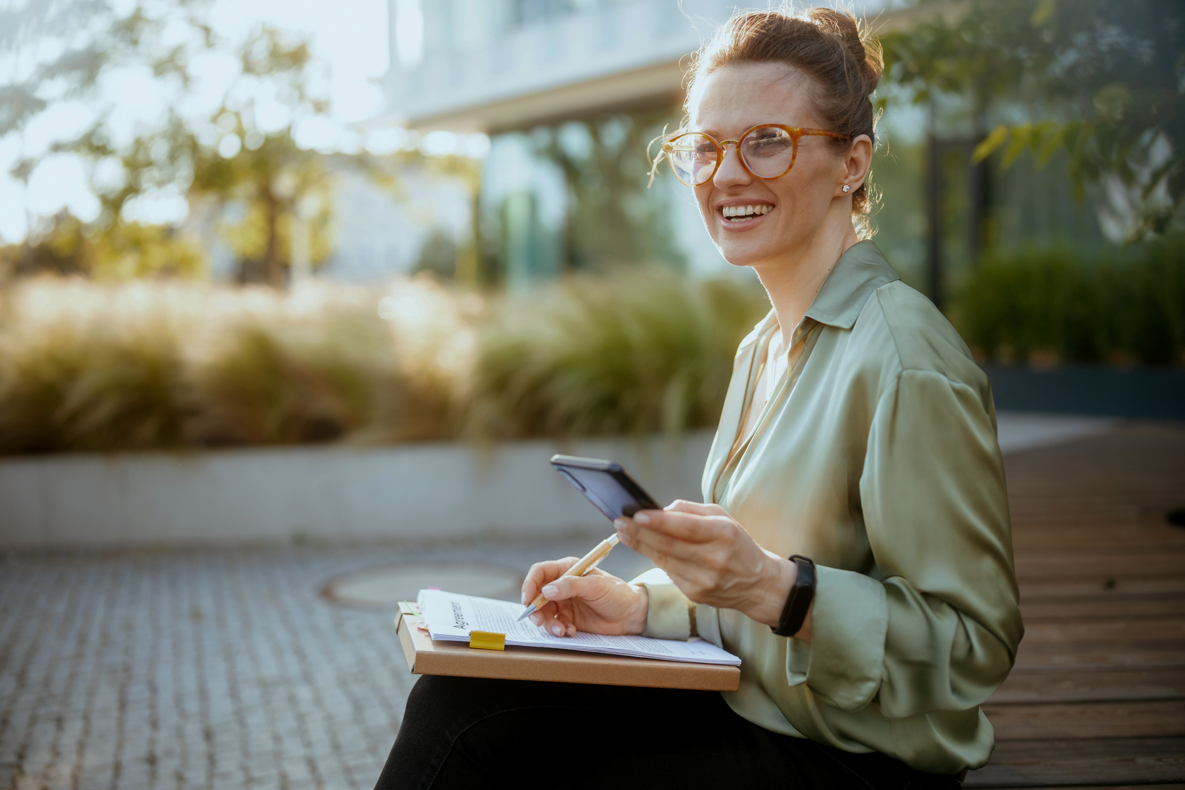 A person sits on a wooden bench, holding a smartphone and a clipboard with notes, surrounded by greenery and soft sunlight.