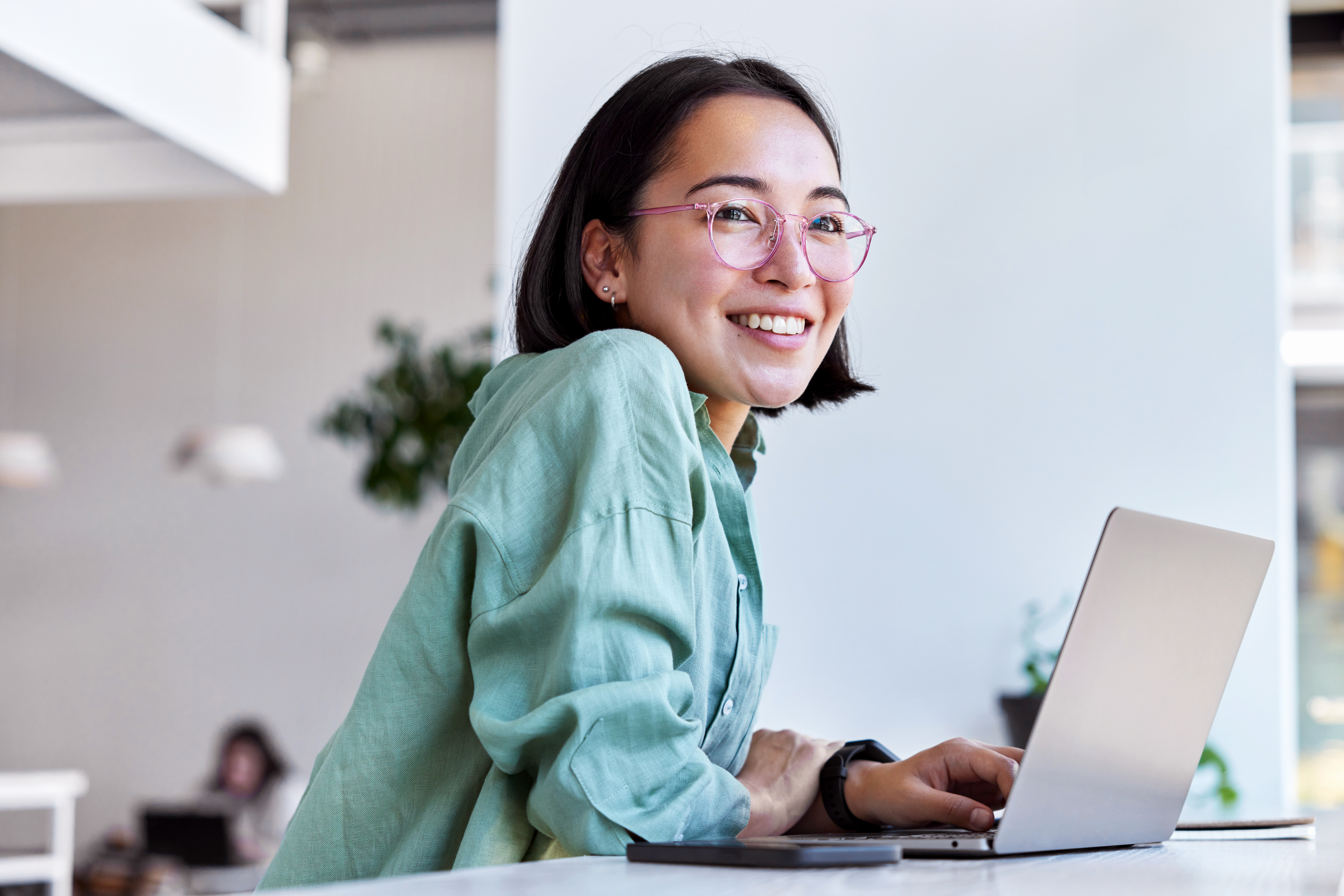 A person in a light green shirt works on a laptop at a table in a modern, bright workspace with plants in the background.