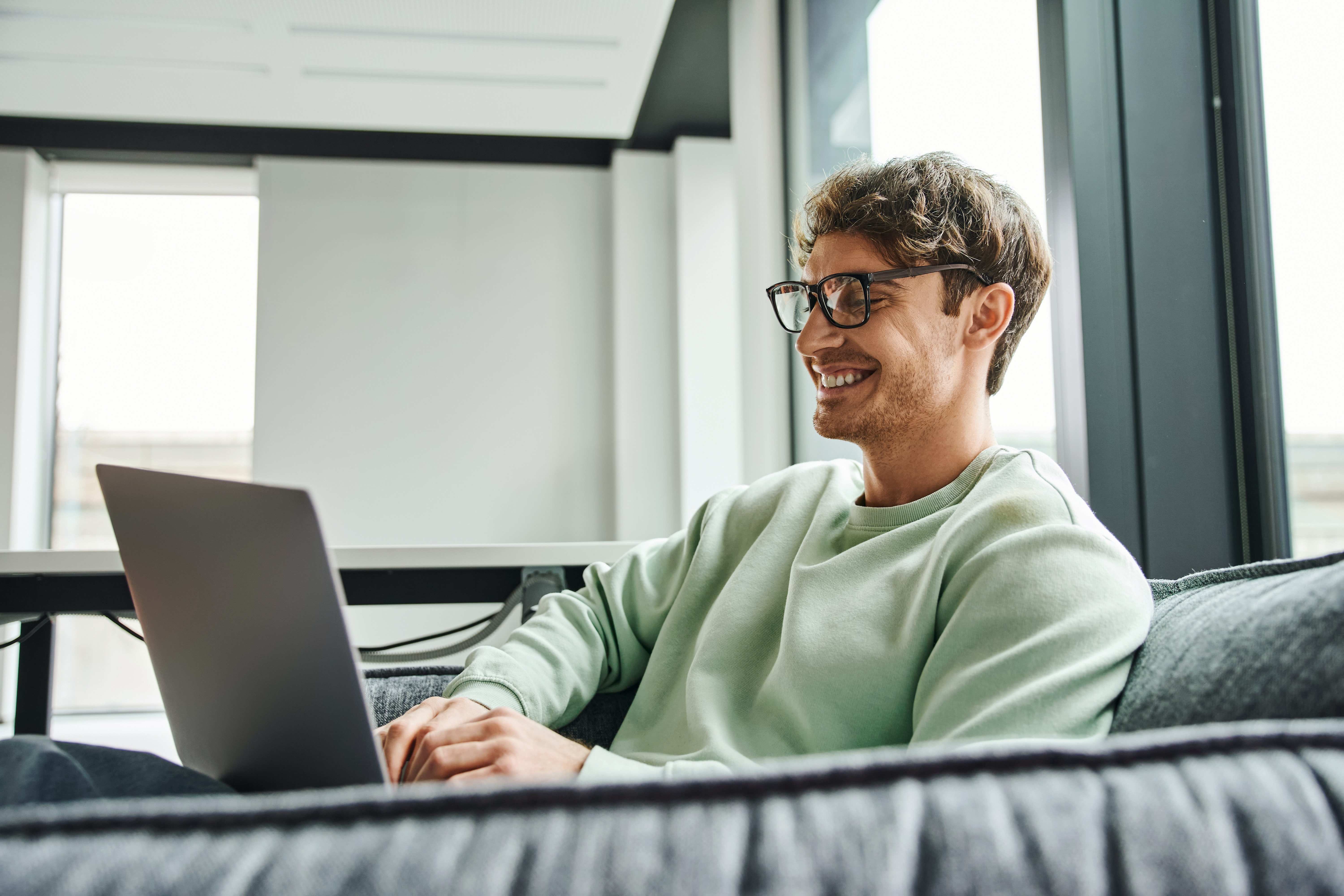 A person in a light green sweater sits casually on a couch, focused on a laptop, with large windows providing natural light.