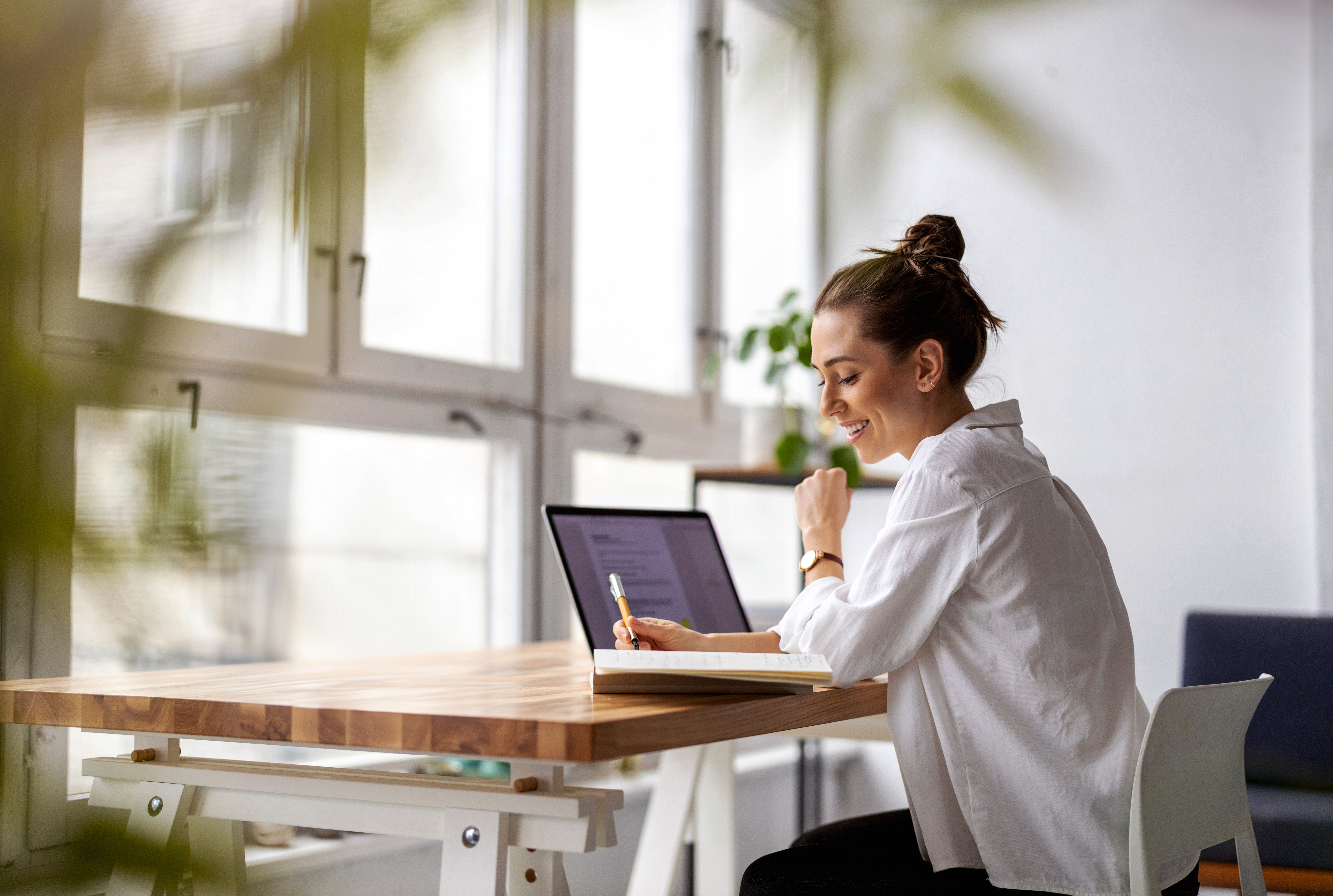 A person sits at a wooden table, working on a laptop, surrounded by plants and minimal decor, in a bright and airy room.