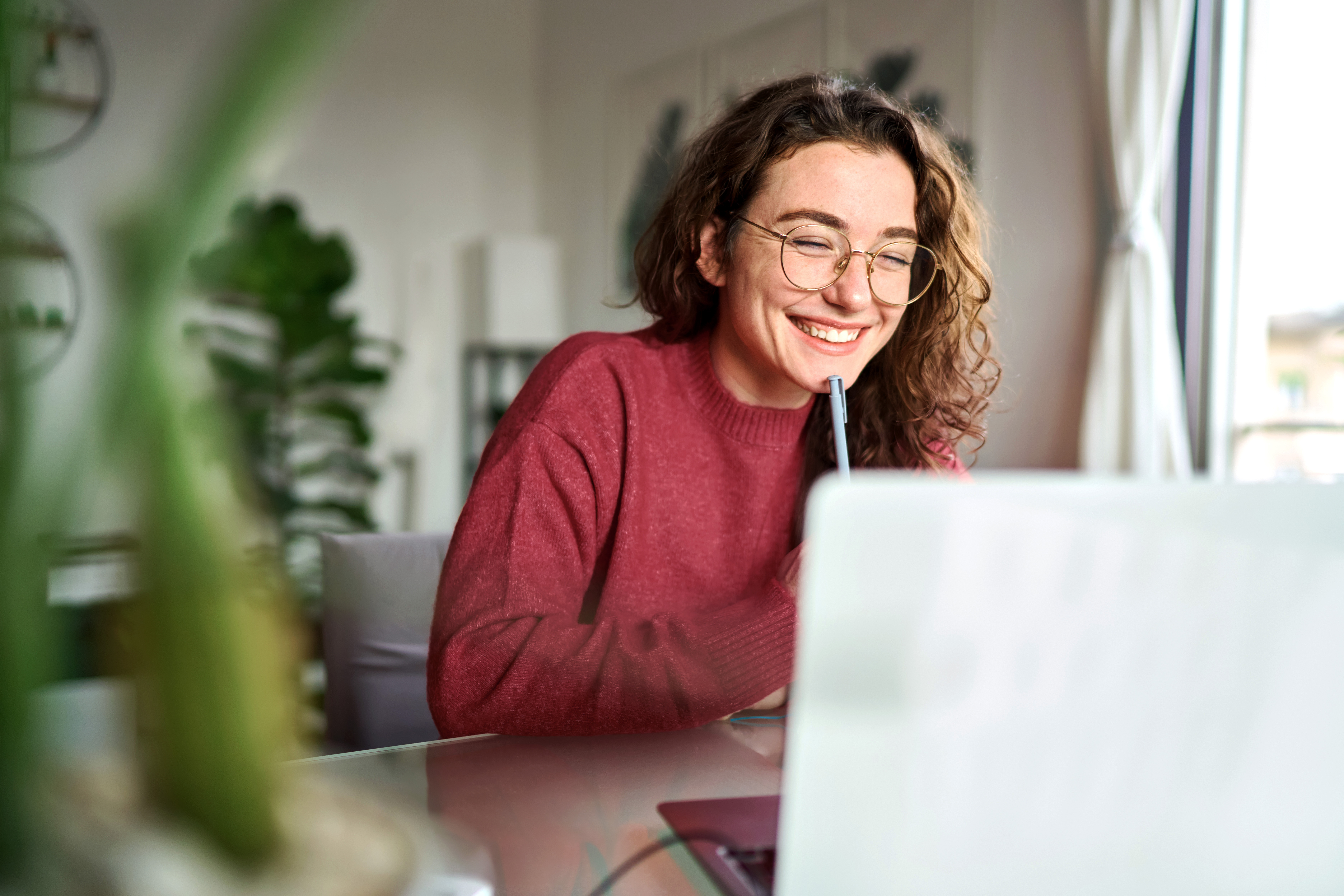 A person in an orange sweater sits at a desk, focused on a laptop, with a smartphone nearby and a plant in the foreground.