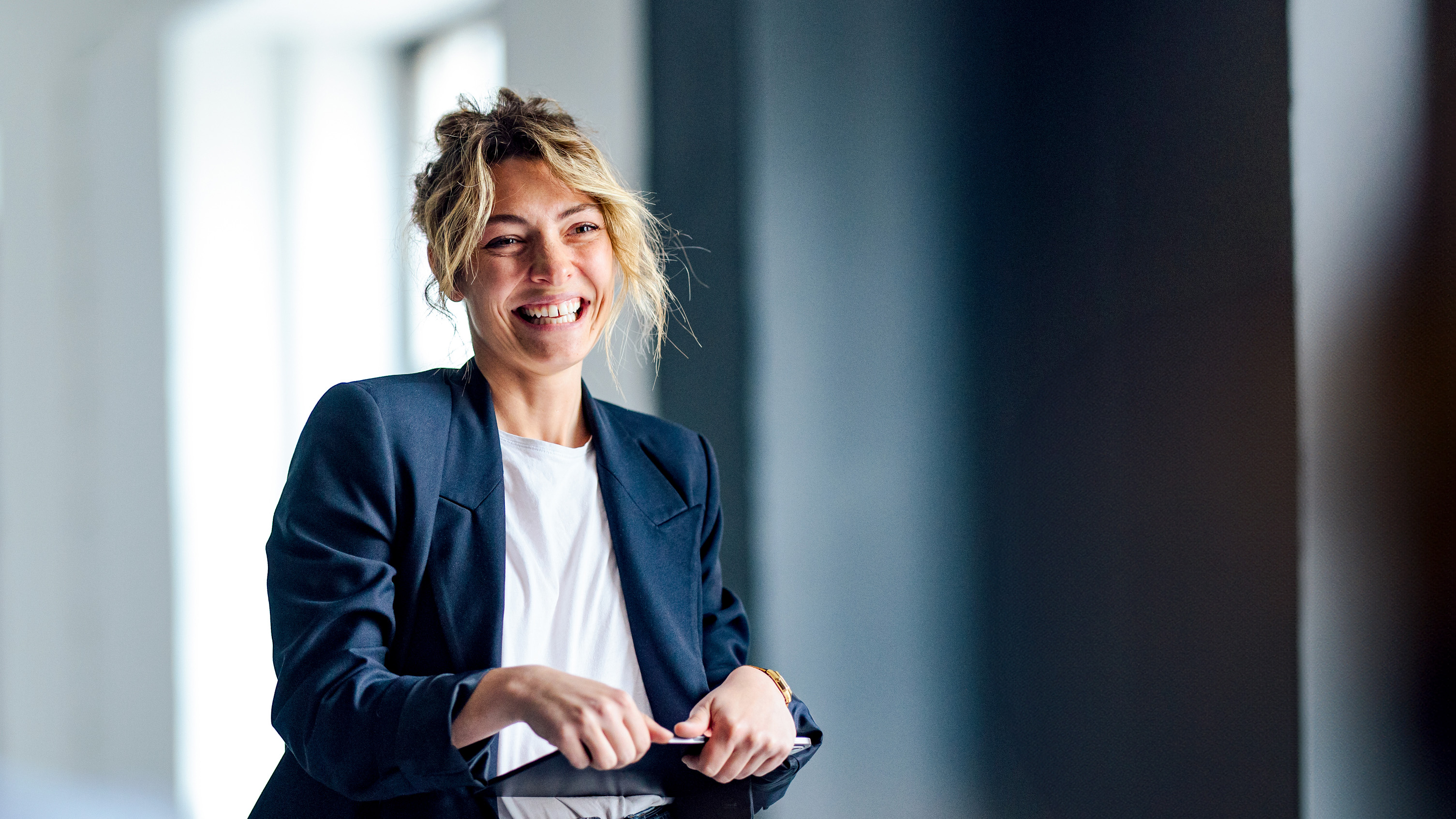 A professional woman wearing a blazer and jeans stands confidently, engaging with an audience in a bright, modern meeting space.