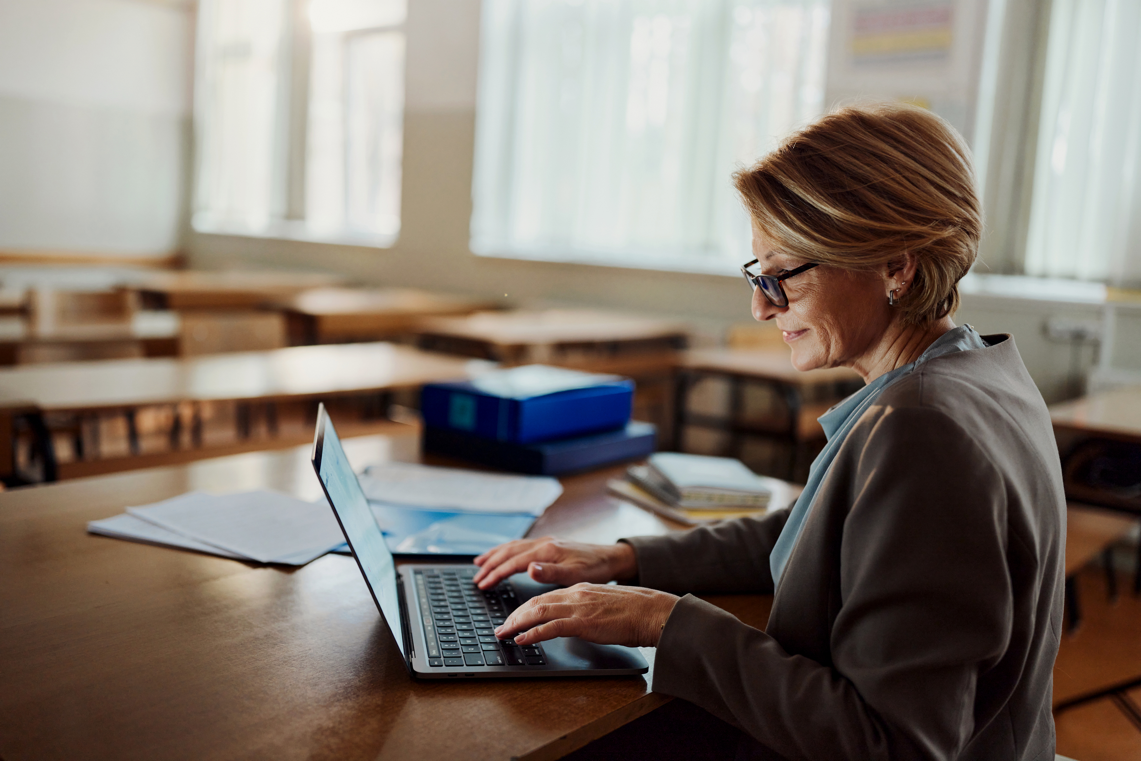 A woman working on a laptop at a wooden table in a classroom, surrounded by papers and office supplies in a bright, airy setting.