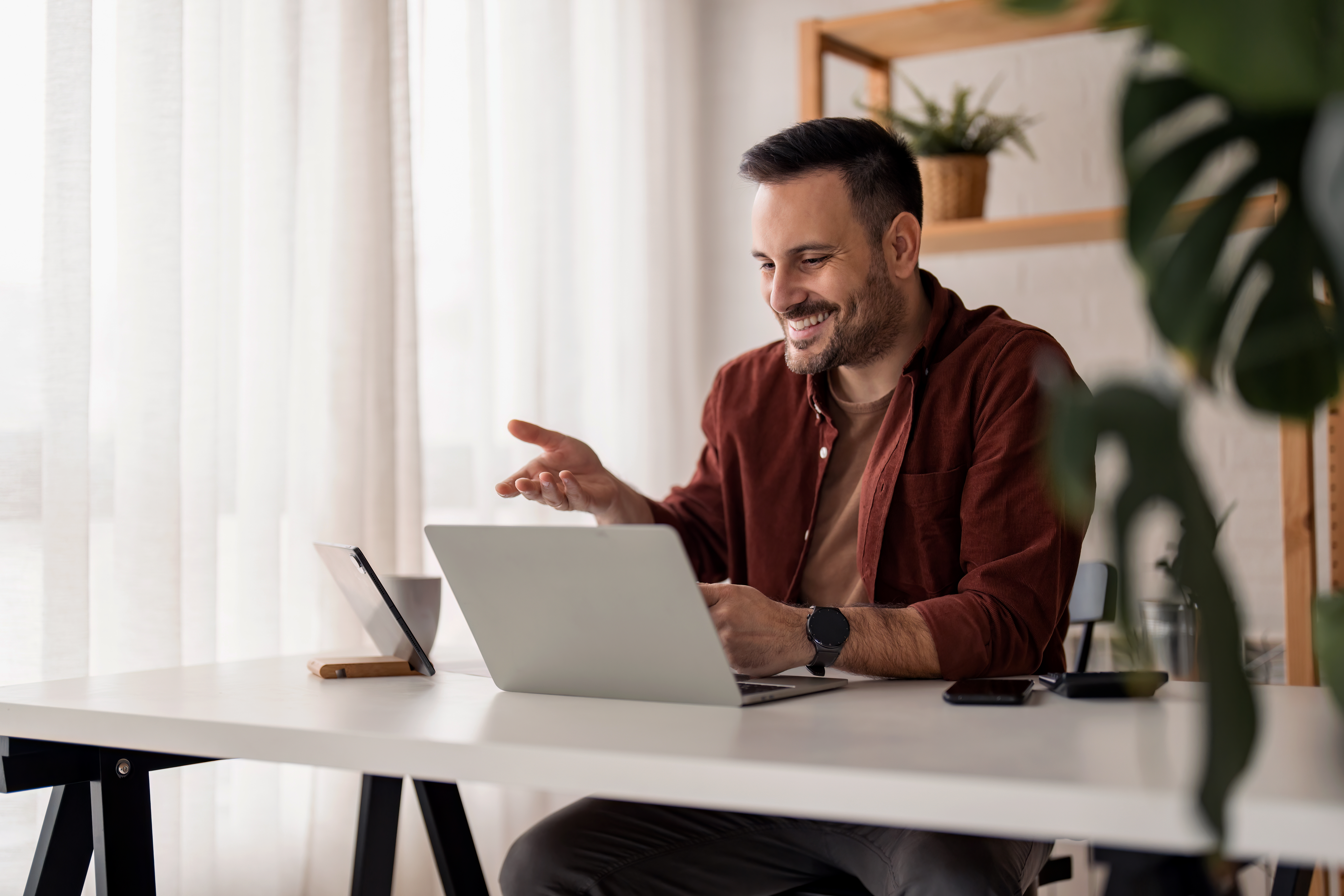 A person in a cozy workspace gestures while engaged in a discussion on their laptop, surrounded by plants and natural light.