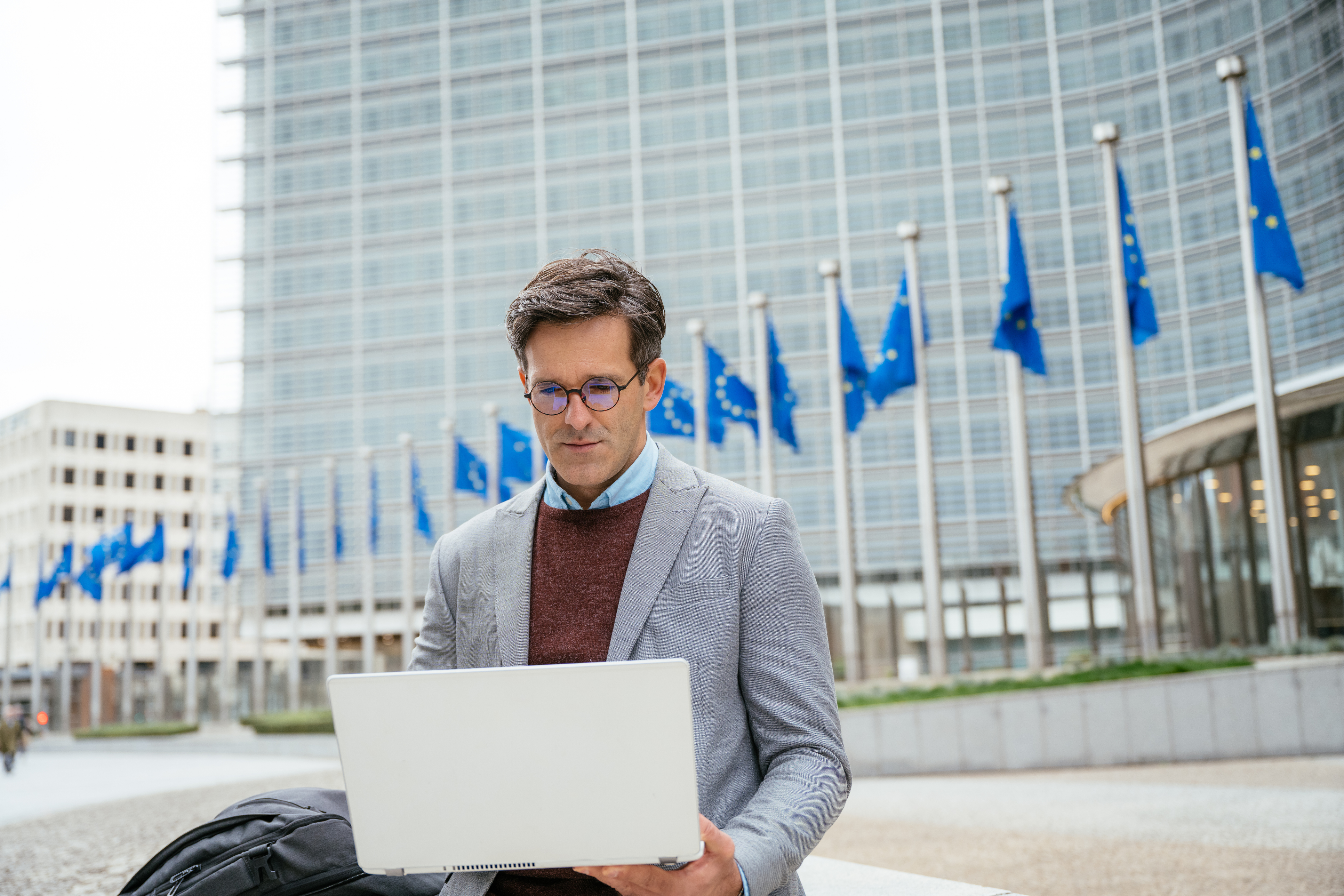 Businessman sitting with laptop in front of business building with European flags
